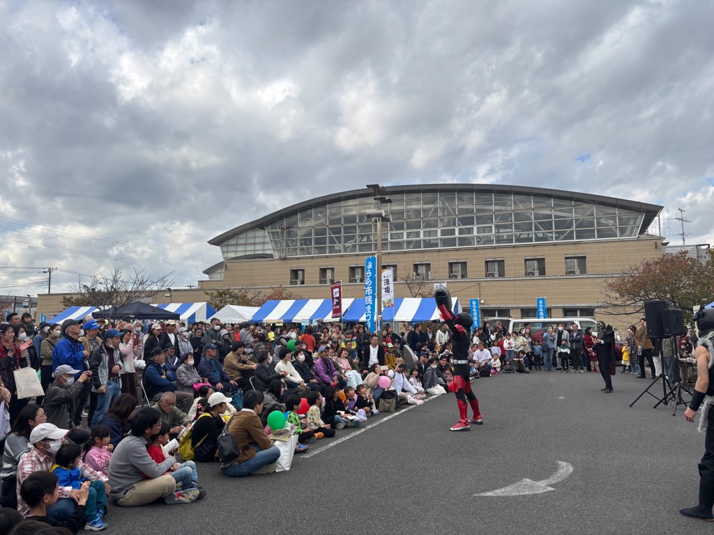 屋外ステージでコントラバスヒーローのヒーローショーを見る大勢の来場者