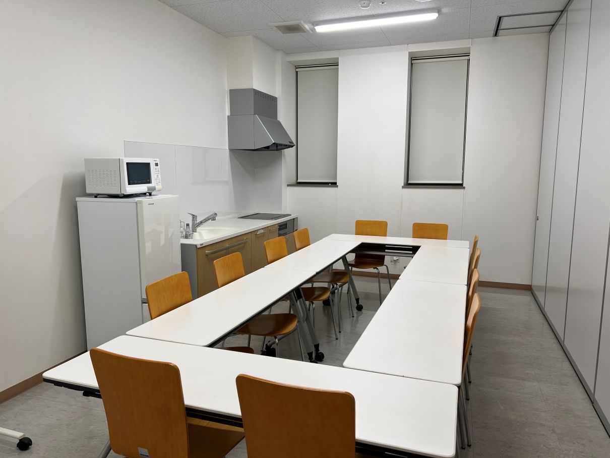 四角に囲んだテーブルに椅子が12個あり冷蔵庫、電子レンジ、流し台が完備された中規模の研修室の写真