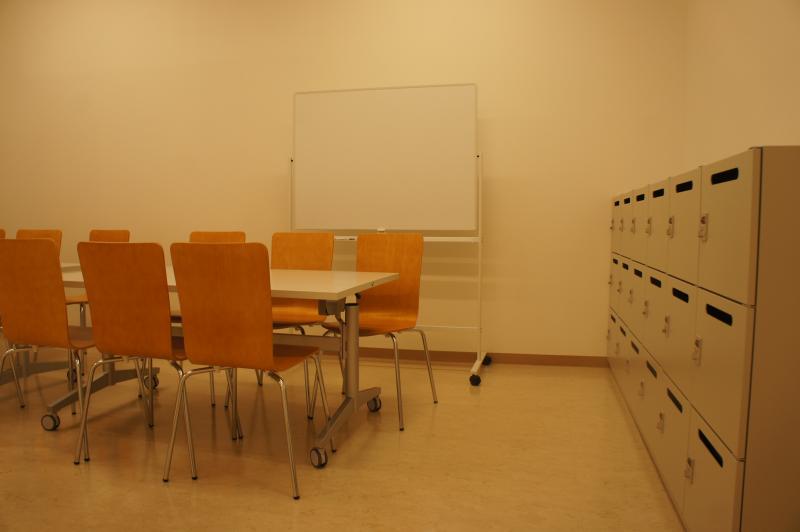 テーブルに椅子が並んでいてロッカーとホワイトボードが置かれている部屋の写真