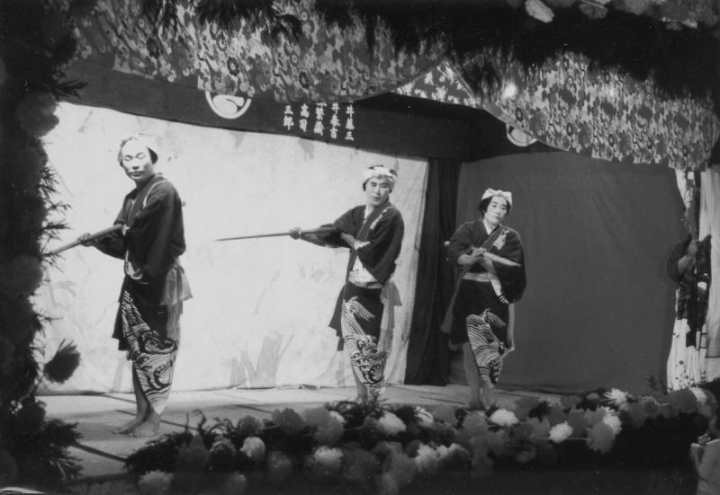 棒を持っている男性3人が舞台で踊っている白黒の写真