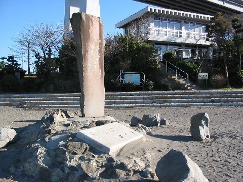 奥の白い建物と木々の間にある階段、手前の砂地に石の台座で立つ石碑の写真
