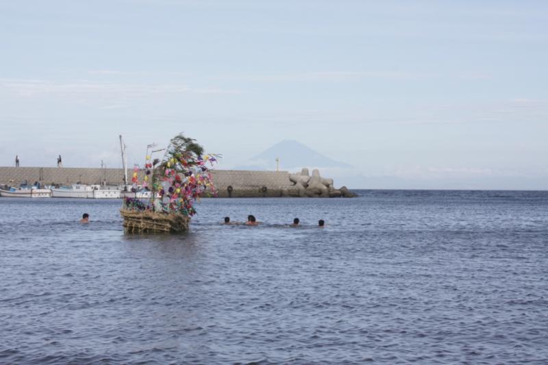 富士山の見える海岸で飾り付けされた船とその周囲を泳ぐ男性たちの写真