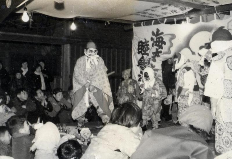 ちょんまげのかつらと面を着けて和服を着た人と和服を着て布を顔に巻いた子供たちを多くの観客が見ている白黒写真