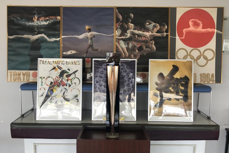 2020年東京オリンピックの聖火リレートーチの後ろに1964年東京オリンピックのポスター大が4枚と2020年東京オリンピックのポスター小が3枚立てかけられている写真