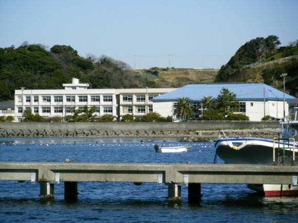 山に囲まれた船が停泊している港の近くに佇む白い校舎と水色の屋根の建物が特徴的な剣崎小学校の写真