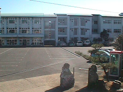 青空の下、たくさんの車が停められる広い駐車場から横長の校舎を写した三崎中学校の写真