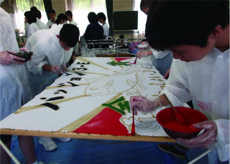 魚のイラストや文字が描かれた旗に、赤い塗料を塗っている男子生徒の写真