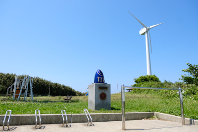 鉄棒やマグロと大根をイメージしたモニュメントに風力発電機のある公園の写真