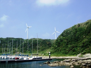 船が並んだ入り江の裏の丘に、白い風車が2本並んでいる写真