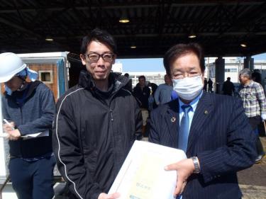 黒いジャージを着た池田水産株式会社専務と、ストライプのスーツを着た三浦市長が記念品を持って並んでいる写真