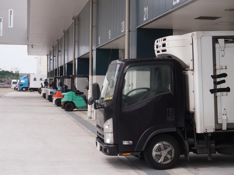 右手前から放射線状に黒に白い荷台のトラック、其並びに緑、赤、灰色、灰色のフォークリフトがそれぞれの車庫に停車している写真