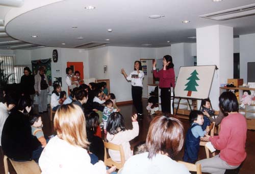 1本の木が描かれたボードの前に立つ2人の女性とその前に座っているたくさんの親子が手拍子をしている写真