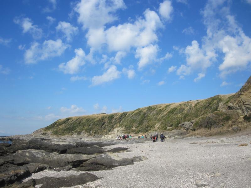 晴れた青空のなか右手に丘が左手に海がある間の砂浜をたくさんの人がリュックを背負って歩いている写真