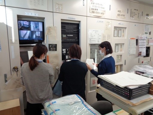3人の女性が監視カメラの映像を映したモニターと黒色の機械が埋め込まれた壁に向かい資料を見ている写真