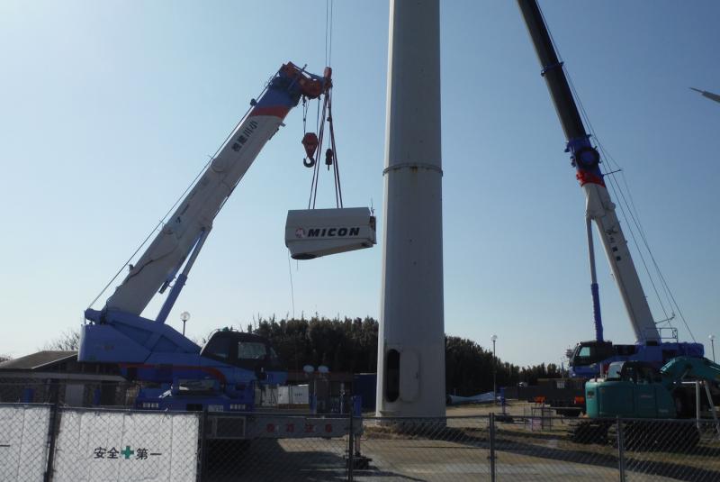 風力発電設備の根本で稼働する2台のクレーン車と吊られる部品の写真