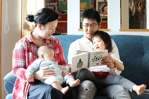 椅子に座りながら子ども二人に絵本を見せている両親の写真