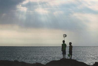 雲の隙間から海面に向かって光が差し込み、岩の上で虫取り網を持って2人の少年が佇んでいる景色の写真