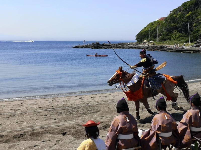 浜辺で馬に乗りながら鎧兜を纏って矢を引いている人物と、それを見ている人物たちの写真