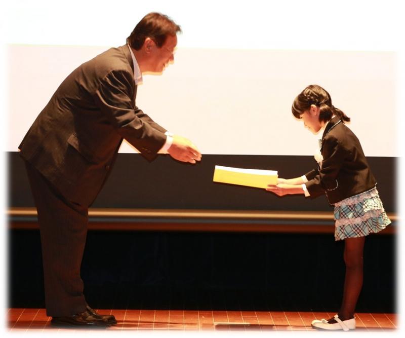 賞状を渡す男性と受け取っている女の子の平和の標語表彰式の写真