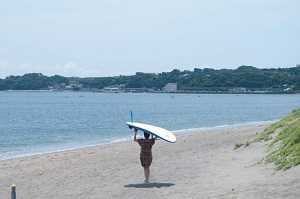 長い砂浜をサーフボードを頭に乗せ歩いている人の写真