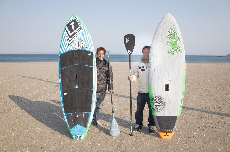 三浦海岸の砂浜で、桑谷さんと男性がサーフボードとパドルを持ち並んでいる写真
